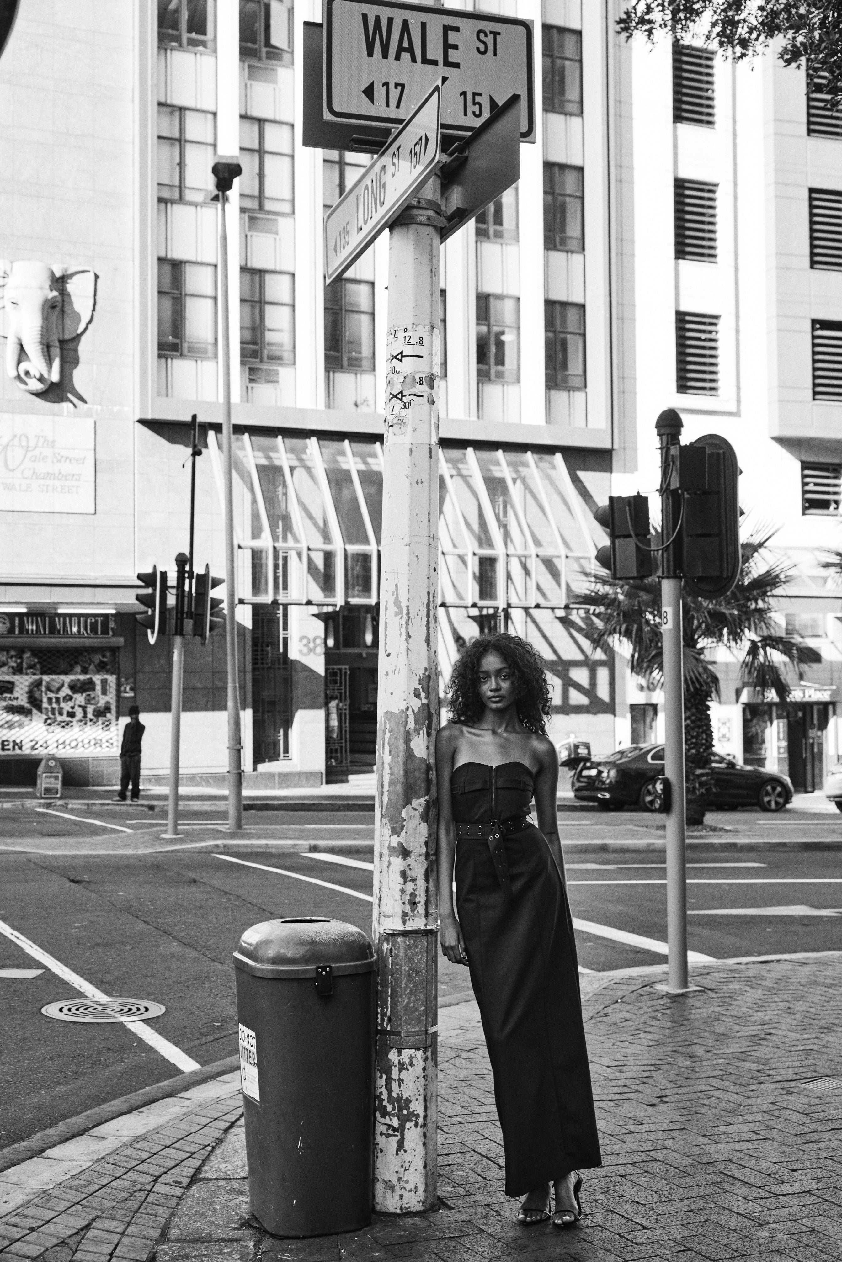 A woman in a long dress and heels standing on a street corner, exuding confidence and style.